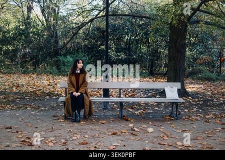 Donna siede sulla panchina nel parco pubblico durante l'autunno mentre guarda a fianco. Gestire l'ansia all'aperto, donna penosa sulla panchina, ritratto dell'umore premuroso Foto Stock