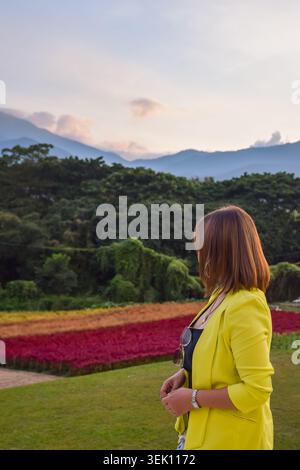 Donna elegante in un blazer giallo che si gode di colorati campi di fiori con paesaggio montano e foresta sullo sfondo durante il tranquillo viaggio al tramonto Foto Stock