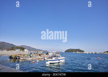 Una tranquilla vista del porto di Namhae Mijo in una giornata di sole, con barche da pesca in acque blu calme, montagne costiere e un chiaro e ampio cielo blu. Foto Stock