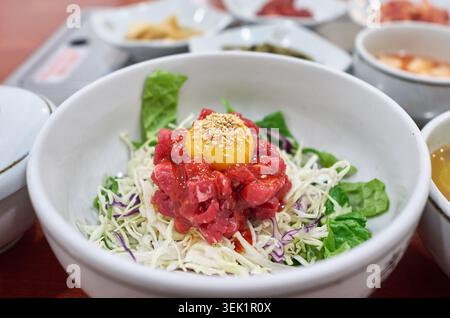Yukhoe Bibimbap coreana con tartare di manzo crudo fresco e tuorlo d'uovo crudo luminoso sulle verdure grattugiate in una ciotola bianca con contorni. Foto Stock