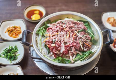 Bulgogi di manzo crudo fresco, spaghetti di vetro e verdure sono disposti in una pentola di metallo, accompagnati da tradizionali contorni coreani e una salsa di tuorli d'uovo. Foto Stock