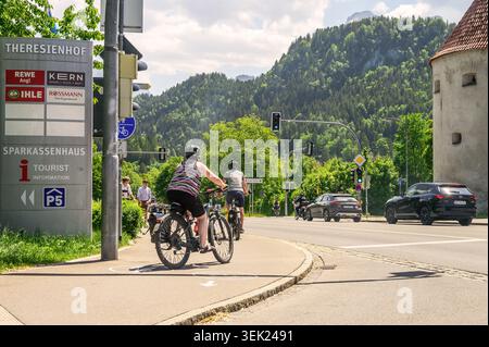Schwangau, Germania - maggio 31 2026: Ciclisti percorrono una pista ciclabile vicino a un cartello Theresienhof con i loghi Rewe e Rossmann e l'Alpe Bavarese Foto Stock