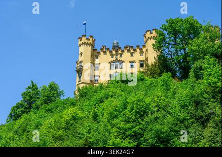 Schwangau, Germania - maggio 31 2026: Facciata del castello di Hohenschwangau circondata da lussureggianti alberi verdi contro un vivace cielo blu. Foto Stock