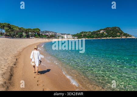 Donna che passeggia lungo la spiaggia sabbiosa di Lloret de Mar con acque turchesi del Mediterraneo Foto Stock