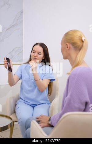 Giovane medico in uniforme consulenza paziente femminile, tenendo smartphone per mostrare qualcosa, e spiegando informazioni sanitarie durante un'app Foto Stock