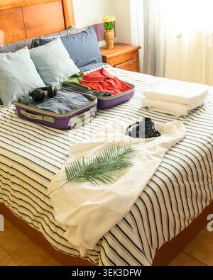 Valigia aperta con vestiti e cuffie, macchina fotografica, asciugamani e foglie di palma su un letto a righe per un viaggio estivo. Foto Stock