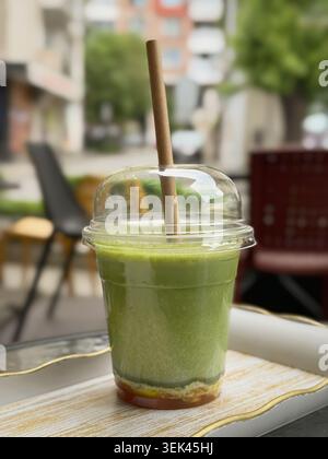 Latte matcha freddo fresco in una tazza da asporto limpida con cannuccia di carta sul tavolo da caffè all'aperto. Sfondo urbano offuscato, stile di vita naturale, bevanda salutare Foto Stock