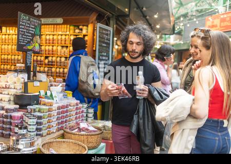 Londra, Regno Unito, 3 maggio 2026, gruppo di persone che guardano un pacchetto di spuntini secchi in un mercato all'aperto con scaffali pieni di barattoli di spezie e. Foto Stock