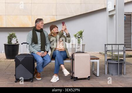 Coppia di mezza età seduta su una panchina di legno al plaza scattando un selfie con smartphone e due valigie Foto Stock
