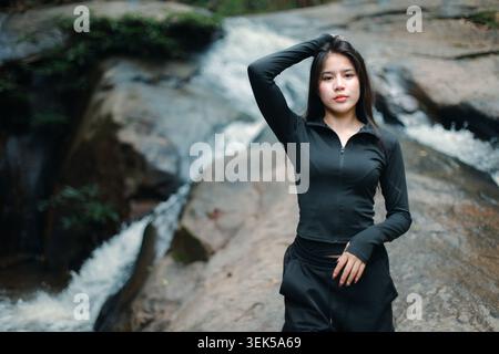 Giovane donna asiatica che posa con sicurezza vicino a una splendida cascata nella giungla tropicale e indossa eleganti abiti atletici con calma e serenità Foto Stock