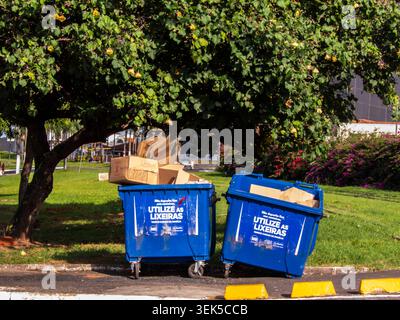 Marilia, SP, Brasile. 16 aprile 2026. Due contenitori blu per rifiuti in plastica riempiti di scatole di cartone per il riciclaggio, situati in un parco pubblico di dow Foto Stock