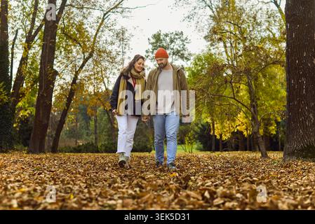 Coppia felice che cammina attraverso il parco autunnale, tenendo per mano le foglie dorate cadute Foto Stock