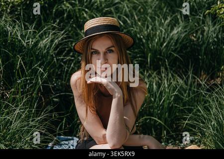 La donna siede tra erba verde alta e indossa un cappello di paglia e poggia il mento sulla mano. Moda estiva sostenibile, vendita al dettaglio eco-compatibile, fibra naturale Foto Stock