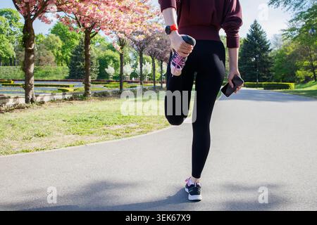 Donna sportiva che si allunga le gambe durante il jogging nel parco cittadino primaverile Foto Stock