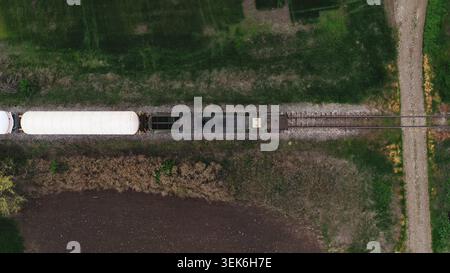 Fotografia aerea dall'alto verso il basso di vagoni merci bianchi che viaggiano su binari ferroviari attraverso terreni agricoli verdi, evidenziando il trasporto rurale e la logistica Foto Stock
