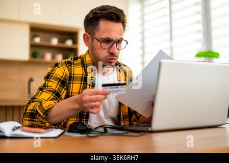 Un giovane ansioso ha una carta di credito mentre lavora a distanza sul suo laptop, sembrando preoccupato e stressato. Foto Stock
