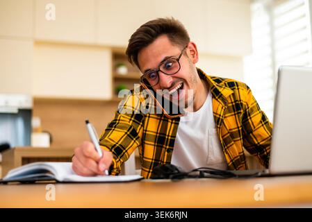 Lavorando da casa, un giovane sta scrivendo idee e promemoria, combinando la ricerca online con il tradizionale prendere appunti. Foto Stock