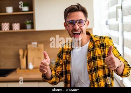 Ritratto di un giovane allegro nella sua cucina. Sta mostrando i pollici in alto. Foto Stock