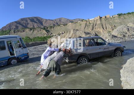 Uomini del villaggio che aiutano a spingere un veicolo fuoristrada attraverso un fiume in un aspro paesaggio di montagna Foto Stock