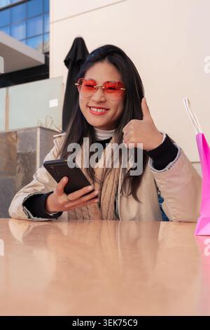 Donna felice che tiene in mano lo smartphone che si regge il pollice dopo aver fatto acquisti con le borse accanto a lei Foto Stock