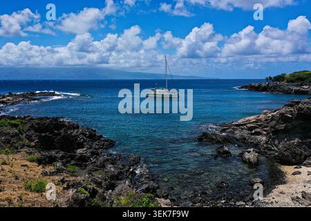 Una vista aerea di una tranquilla baia a Maui, Hawaii, caratterizzata da un catamarano ancorato sulle vibranti acque turchesi. Intorno alla scena c'è la roccia lavica Foto Stock