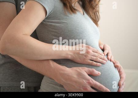 Donna incinta con partner che mette le mani sopra la pancia in un momento intimo condiviso. Aspettarsi che madre e marito abbracciino da fianco in studio con background neutrale. Mamma che festeggerà in comune Foto Stock