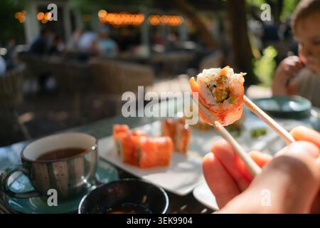 Primo piano di sushi di salmone fresco tenuto con bacchette, caratterizzato da un autentico ambiente da ristorante giapponese con un pasto in sottofondo per i bambini. Foto Stock
