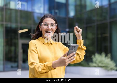 Giovane donna in occhiali che alza il pugno in un gesto di successo ed eccitazione, reagendo alle buone notizie sul suo smartphone all'aperto di fronte a un moderno edificio di uffici Foto Stock