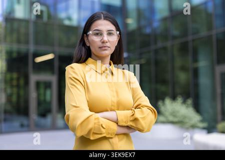 Donna d'affari con le braccia incrociate, con occhiali trasparenti e una camicia gialla, che rappresenta professionalità e leadership in un ambiente urbano aziendale Foto Stock