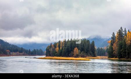 Fiume che si snoda attraverso una tranquilla natura selvaggia con alberi autunnali e montagne nuvolose Foto Stock