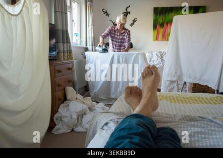 divertente immagine di un uomo sdraiato a letto con i piedi incrociati a guardare la moglie, il partner che lavora a stirare circondato da un'asciugatura in lavatrice, Foto Stock