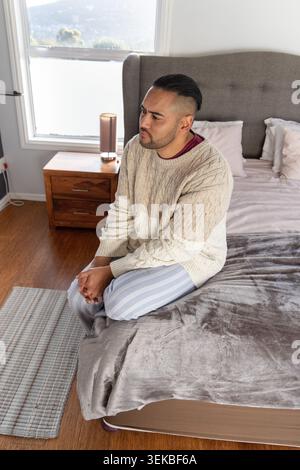 Uomo di trent'anni seduto sul letto con coperta in camera da letto, con maglione e pigiama, mani che stringono Foto Stock