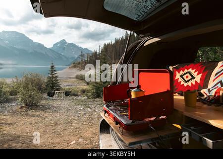 vista sul retro di un camper con una mensola con una stufa rossa da campo in estate con la luce del mattino e montagne sullo sfondo Foto Stock