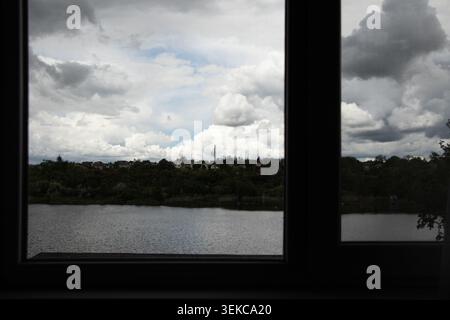 Una vista da una finestra di un fiume, alberi ed edifici residenziali sotto spesse nuvole di pioggia per temi quali il tempo, la pioggia, la finestra, la natura e il paesaggio Foto Stock