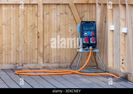 Scatola di distribuzione elettrica portatile con supporti per cavi di alimentazione arancioni su un pavimento in legno accanto a pareti in legno non finite. Foto Stock