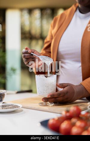 Primo piano, vlogger di cibo, miscelazione del budino di chia, ingredienti nel recipiente, semi di chia nutrienti, colazione allo yogurt, ricetta per preparare i pasti. Influencer nero che presenta un'idea sana ricca di proteine. Foto Stock