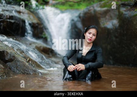 Bella donna asiatica che indossa un costume da bagno nero seduto in acqua, si sente rilassata e serena, con splendide cascate e grandi rocce bagnate sul retro Foto Stock