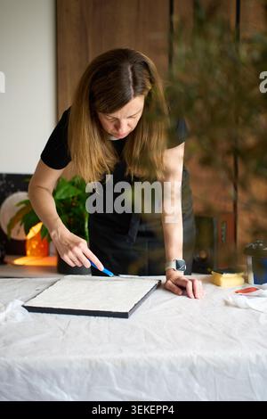 La donna in grembiule nero crea opere d'arte su tela a casa, diffondendo pasta bianca con un coltello blu su un tavolo rivestito di tessuto. Concetto di arredamento per la casa fai da te e hobby creativo Foto Stock