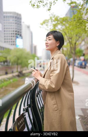 Una giovane donna sta accanto alle ringhiere della città, guardando in lontananza. Indossa un cappotto beige, tiene il telefono, sembra premurosa. Verde urbano e alti edifici Foto Stock