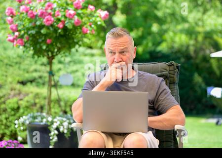 Uomo maturo che lavora con cura su un notebook mentre si rilassa all'aperto in uno splendido giardino verde. Immagine naturale dello stile di vita che rappresenta il lavoro remoto, onl Foto Stock