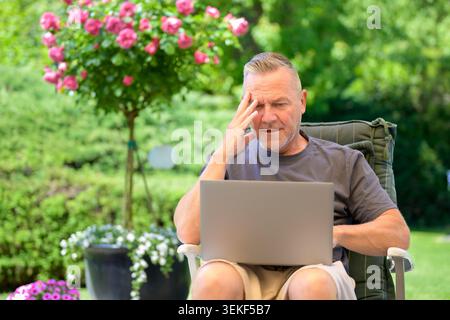 Uomo maturo seduto all'aperto in un bellissimo giardino mentre lavora su un notebook e sembra stressato o preoccupato. Concetto di lavoro remoto, tecnologia probl Foto Stock