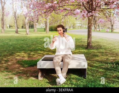 un freelance ama lavorare a distanza in un parco primaverile sotto i fiori rosa. L'uomo si siede con il suo portatile, parla al telefono e tiene il caffè. Alta qualita' Foto Stock