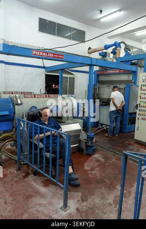 Lavoratore che osserva il processo di pressatura in un oleificio d'oliva. Olio vergine fresco che scorre dai moderni macchinari ai contenitori. Simboleggia la tradizione Foto Stock