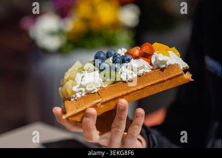 Uno shot ravvicinato della mano di una persona che tiene in mano un waffle belga dorato carico di panna montata, mirtilli, fragole, ananas e fette di kiwi Foto Stock