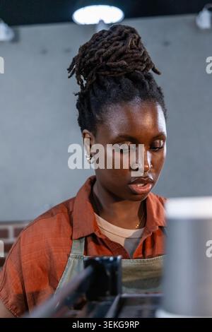 Donna africana adulta che lavora dietro la macchina per espresso commerciale al bancone del bar con grembiule verde Foto Stock
