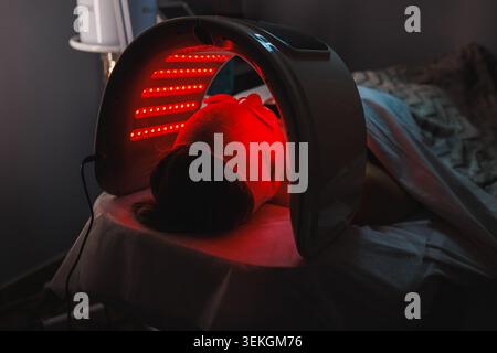 Donna caucasica è sdraiata su un tavolo di trattamento durante il suo appuntamento cosmetologico. Sta ricevendo una terapia a luce rossa per la cura del viso. L'impostazione è A. Foto Stock