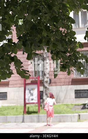 Donna con una gonna a pois rossa e camicia bianca che cammina su un marciapiede, passando davanti a un tabellone rosso all'ombra di un albero verde, catturando un luminoso Foto Stock