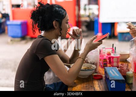 Giovane donna asiatica che usa il telefono al banco dello Street food, gustando un pasto informale Foto Stock