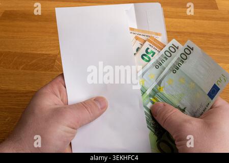 Primo piano di mani maschili con una busta di carta bianca piena di banconote in euro su un tavolo di legno. Concetto di corruzione, corruzione, transazione illegale, b Foto Stock