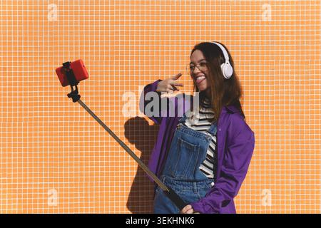 Giovane donna sorridente, indossa cuffie e occhiali, scatta un selfie all'aperto con una parete arancione Foto Stock